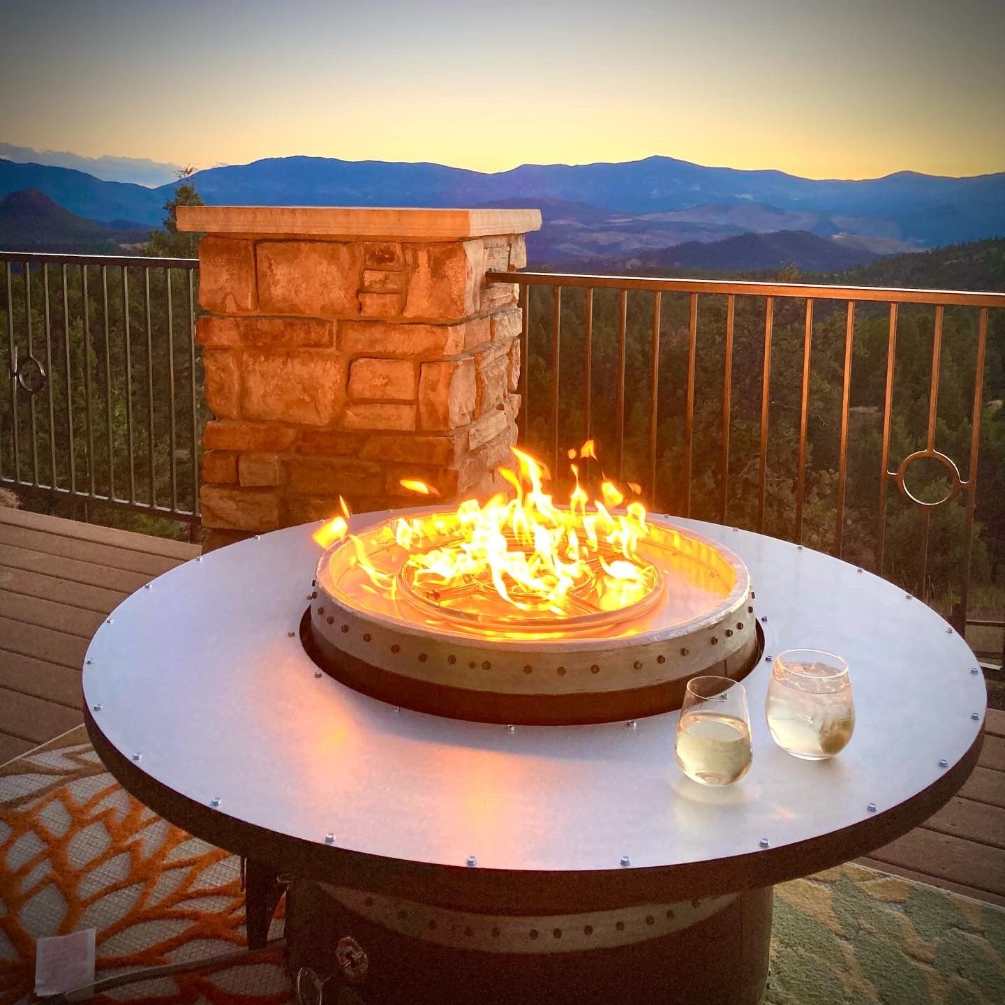 Ring-top fire pit table with a flame and two drinks on a deck at sunset, mountains behind
