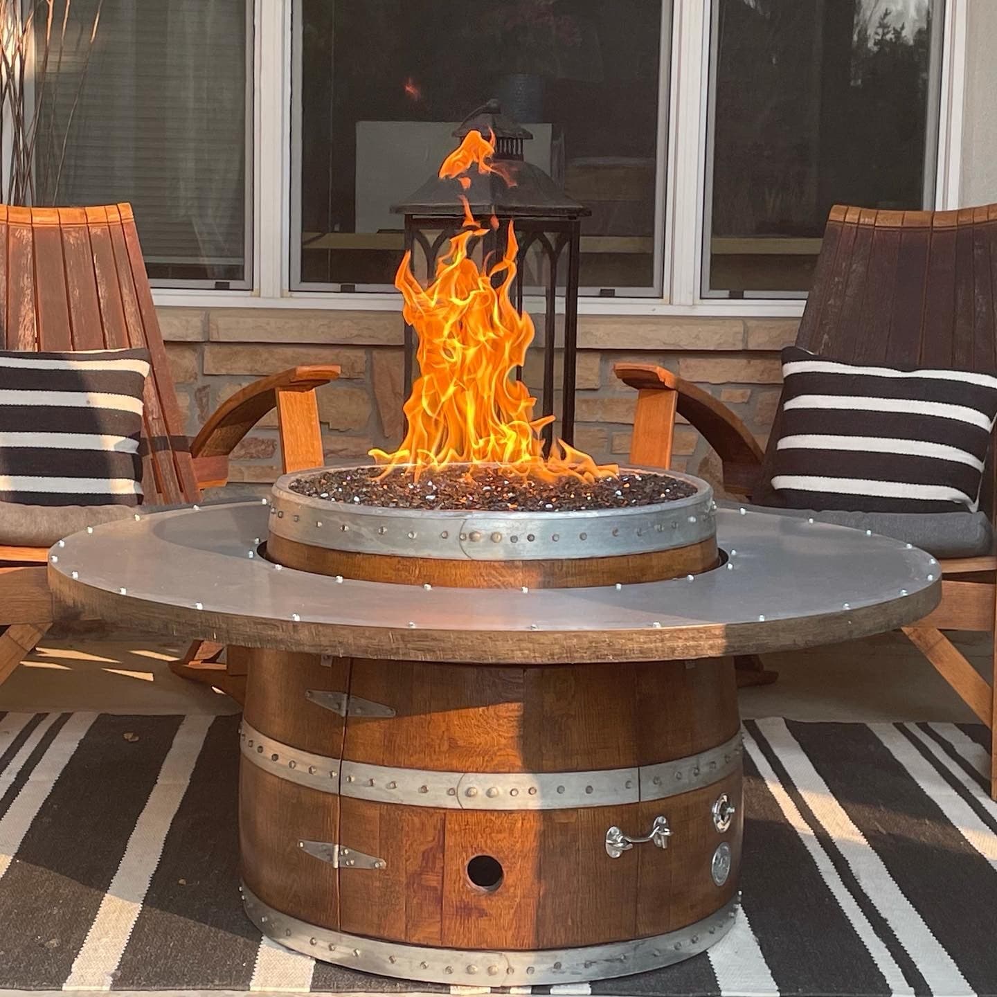 Low barrel fire pit with a ring tabletop and flame between wooden chairs on a striped rug