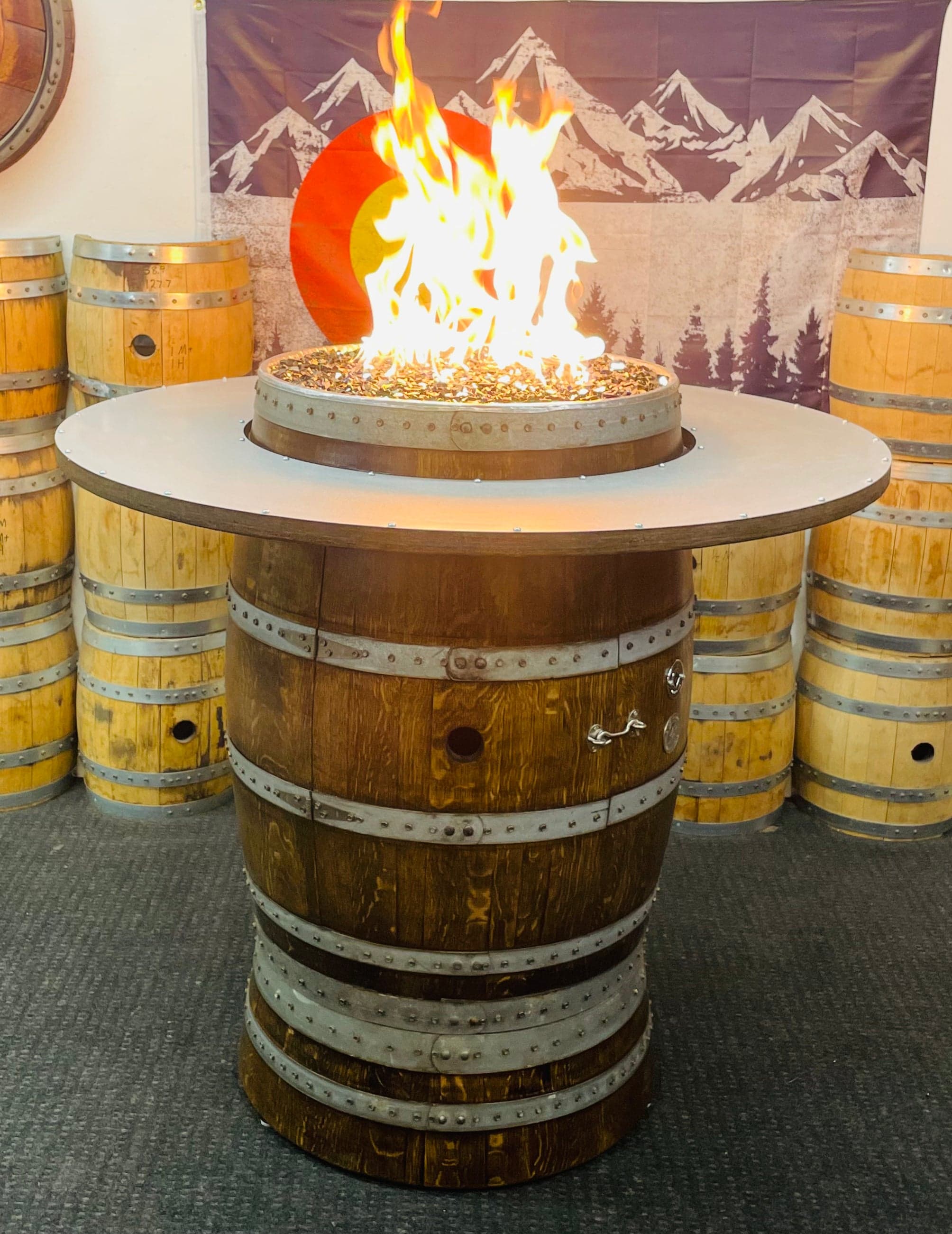 Tall barrel fire pit table with a big flame in front of stacked barrels
