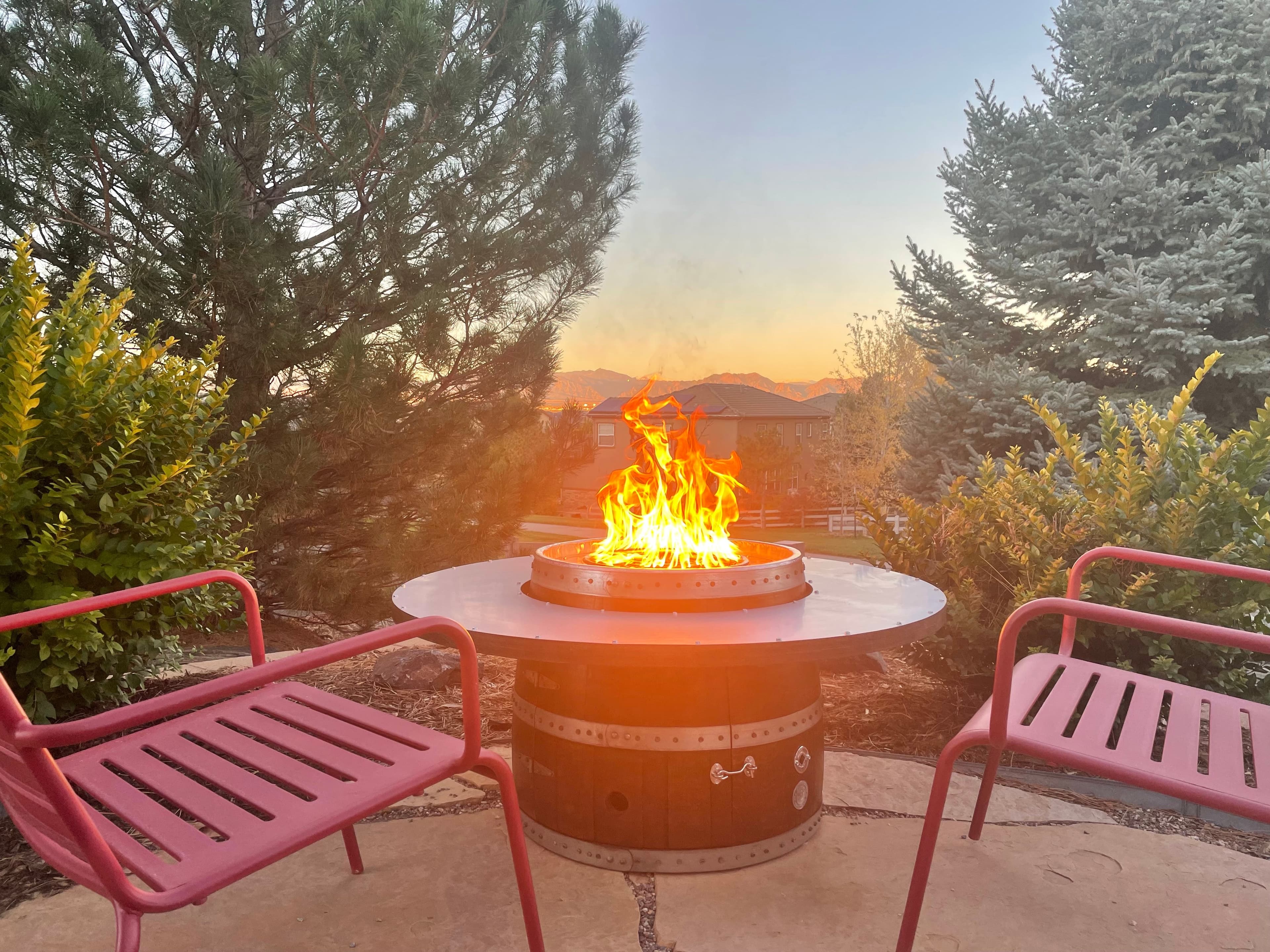 Wine barrel fire pit with a tall flame between two red chairs at sunset, mountains on the horizon