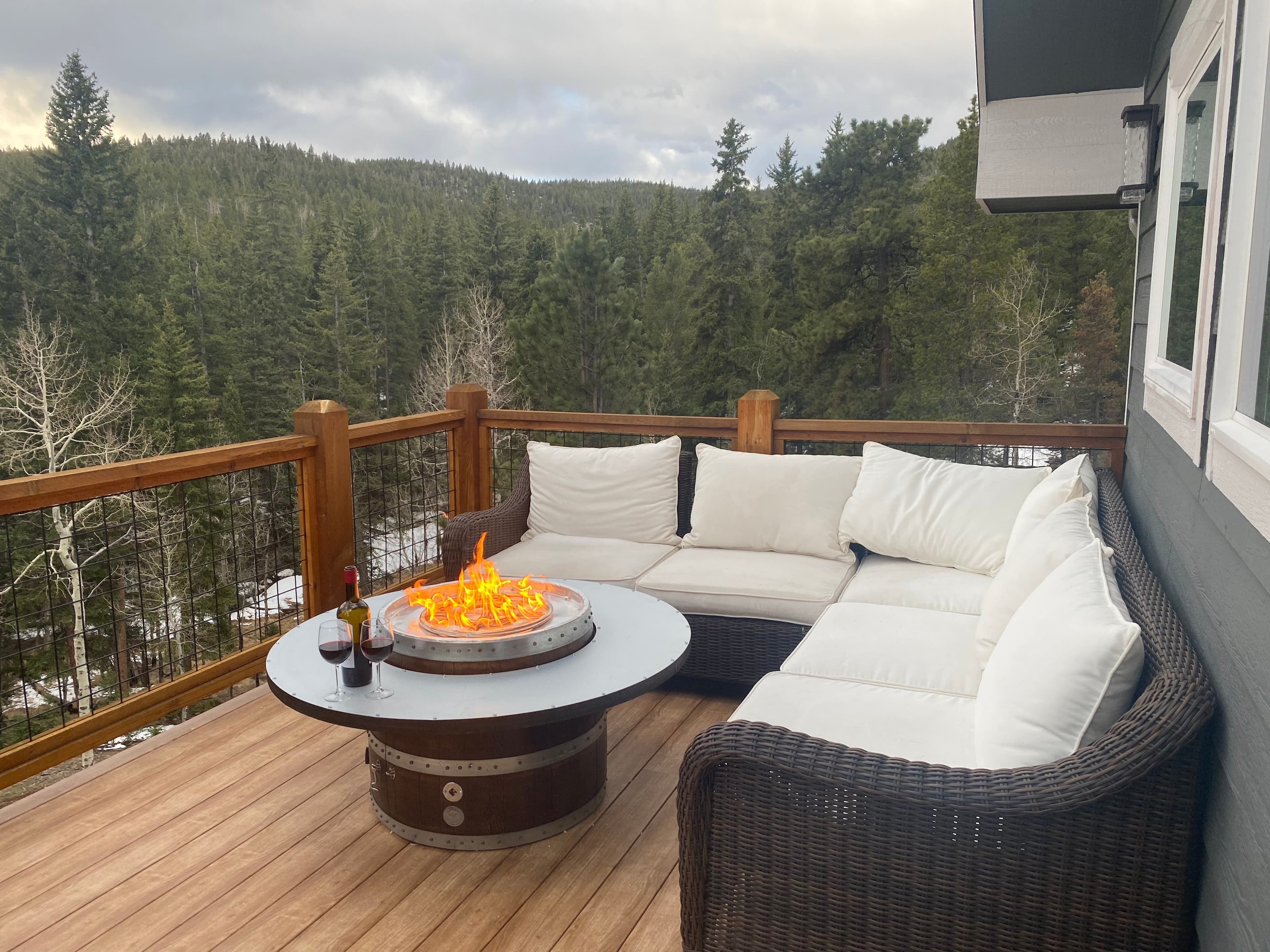 Barrel fire pit table on a mountain deck beside a sectional sofa, forest beyond
